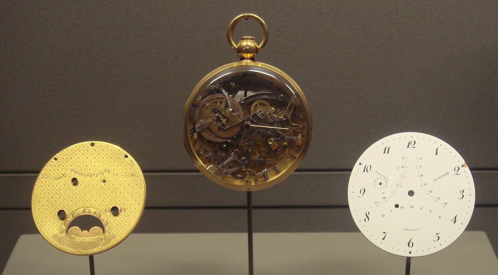 Drei Ausstellungsstücke nebeneinander: eine goldene Werkplatte, eine Taschenuhr mit sichtbarem Räderwerk und ein weisses Emailzifferblatt mit der Aufschrift Breguet