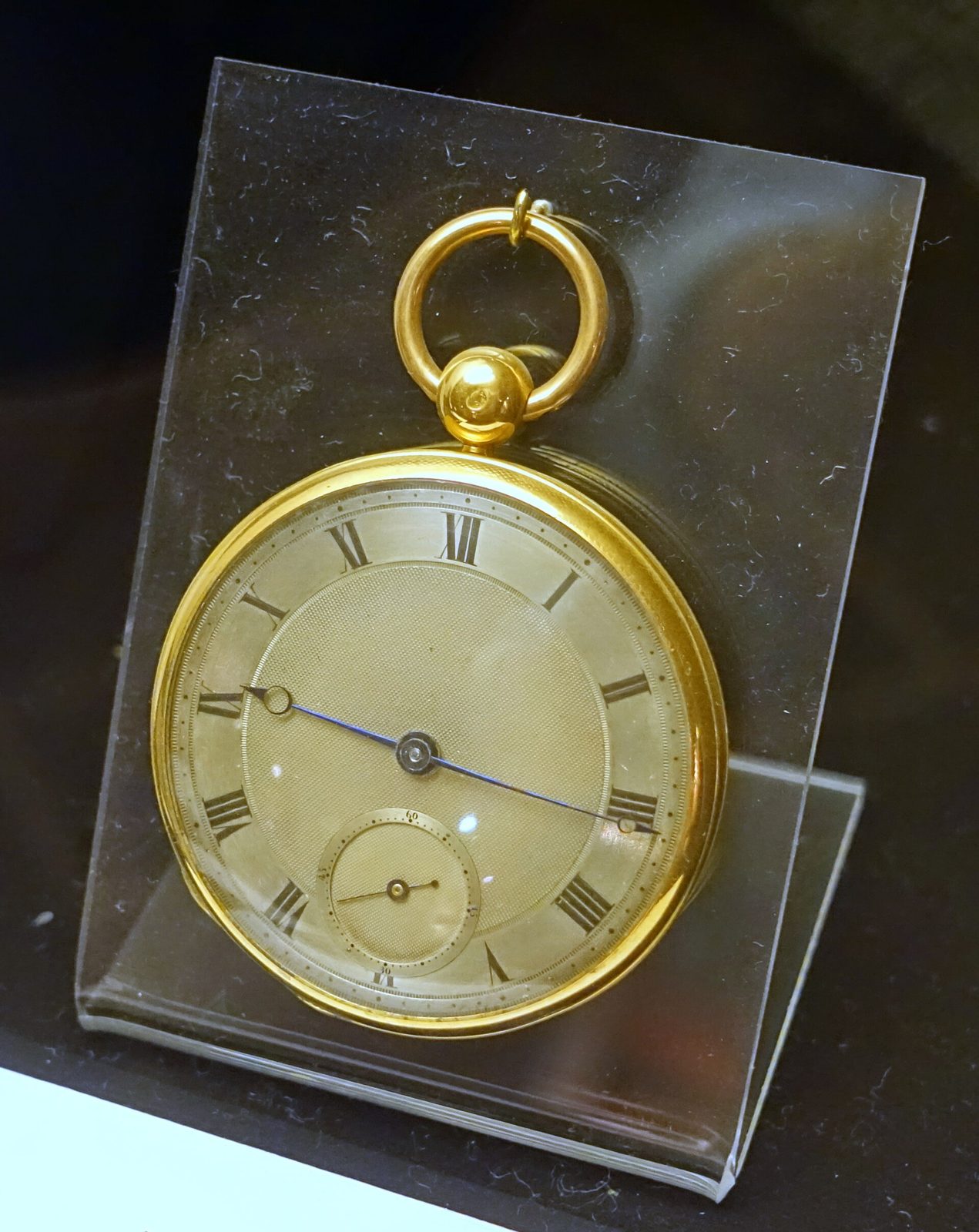 Goldene Taschenuhr mit silbrig guillochiertem Zifferblatt, römischen Ziffern, kleiner Sekunde und blauen Stahlzeigern auf einem durchsichtigen Ständer