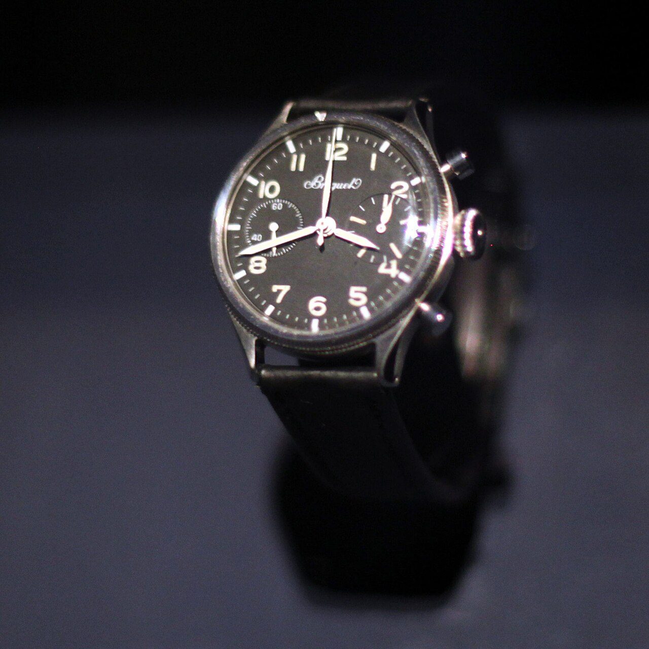 Chronograph mit schwarzem Zifferblatt, Leuchtziffern, zwei Hilfszifferblättern und zwei Drückern an einem schwarzen Band, vor dunklem Hintergrund
