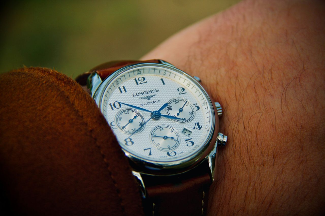 Silberner Automatik-Chronograph mit weissem Zifferblatt und braunem Lederband an einem Handgelenk vor dunklem Hintergrund