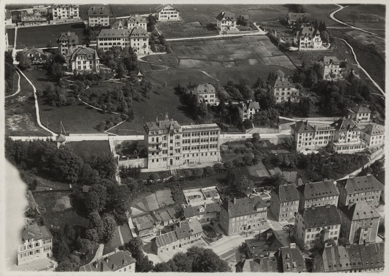 Schwarzweisses Luftbild von Le Locle mit einem grossen Fabrikgebäude zwischen Wohnhäusern und Gärten; im Archiv der ETH-Bibliothek als Uhrenfabrik H. Moser & Cie. verzeichnet