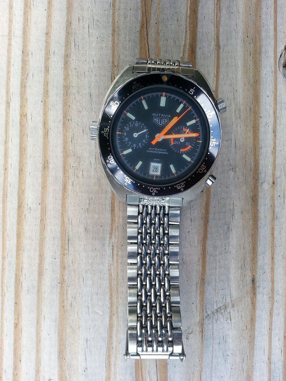 Vintage-Chronograph Heuer Autavia mit schwarzer Drehlünette, dunklem Zifferblatt, orangen Zeigern und Datumsfenster an einem Edelstahl-Gliederband