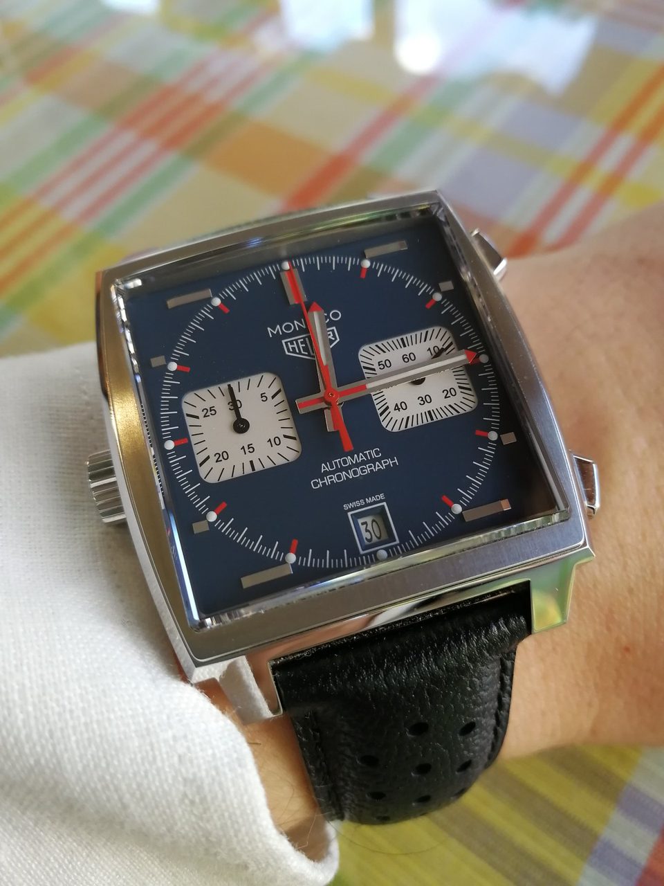 Quadratischer Chronograph Heuer Monaco mit blauem Zifferblatt, zwei weissen Hilfszifferblättern und roten Zeigerspitzen, am Handgelenk über einer karierten Tischdecke