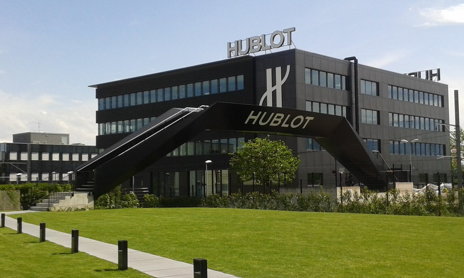Dunkles Bürogebäude mit dem Schriftzug Hublot, davor eine Fussgängerbrücke mit Hublot-Schriftzug und eine Rasenfläche