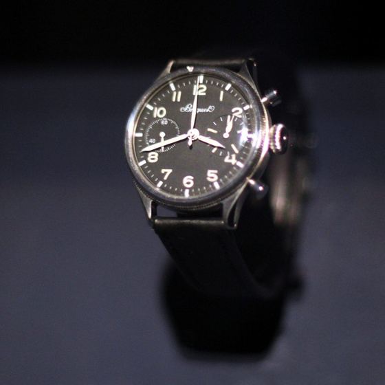 Chronograph mit schwarzem Zifferblatt, Leuchtziffern, zwei Hilfszifferblättern und zwei Drückern an einem schwarzen Band, vor dunklem Hintergrund