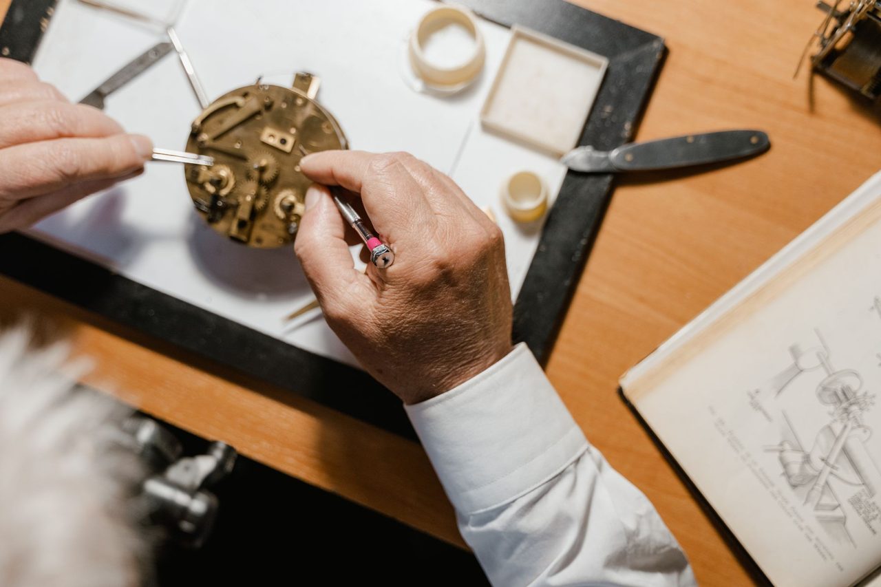 Draufsicht auf einen Werktisch mit Uhrwerk, Werkzeugen und Unterlage, zu sehen sind Hände und Ärmel, kein Gesicht