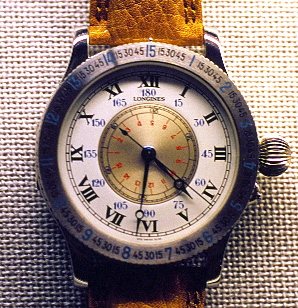 Longines-Armbanduhr mit weissem Zifferblatt, römischen Ziffern, Gradzahlen von 15 bis 180 auf dem inneren Ring, goldfarbener Sekundenscheibe in der Mitte und drehbarer Lünette mit blauen Zahlen an einem gelbbraunen Lederband