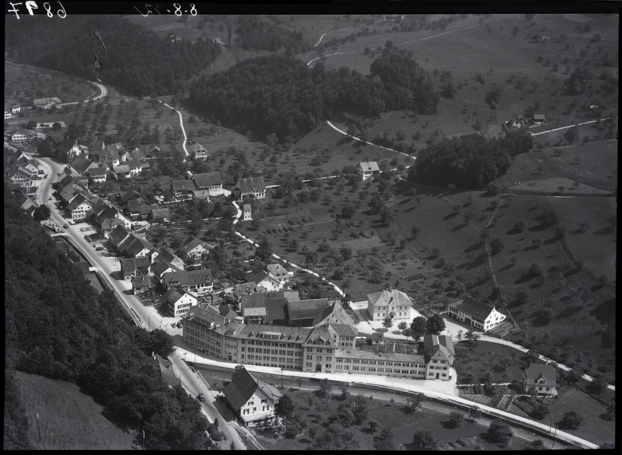 Schwarzweisse Luftaufnahme von Hölstein im Tal mit Häusern, Strassen und einem grossen, langgestreckten Fabrikgebäude im Vordergrund