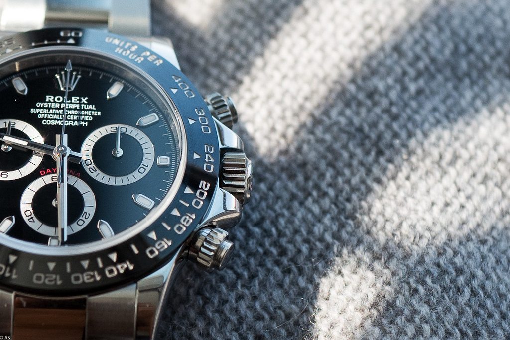 Ausschnitt einer Stahl-Daytona mit schwarzer Keramiklünette, Tachymeter-Skala, Krone und beiden Chronographen-Drückern auf grauem Wollstoff.