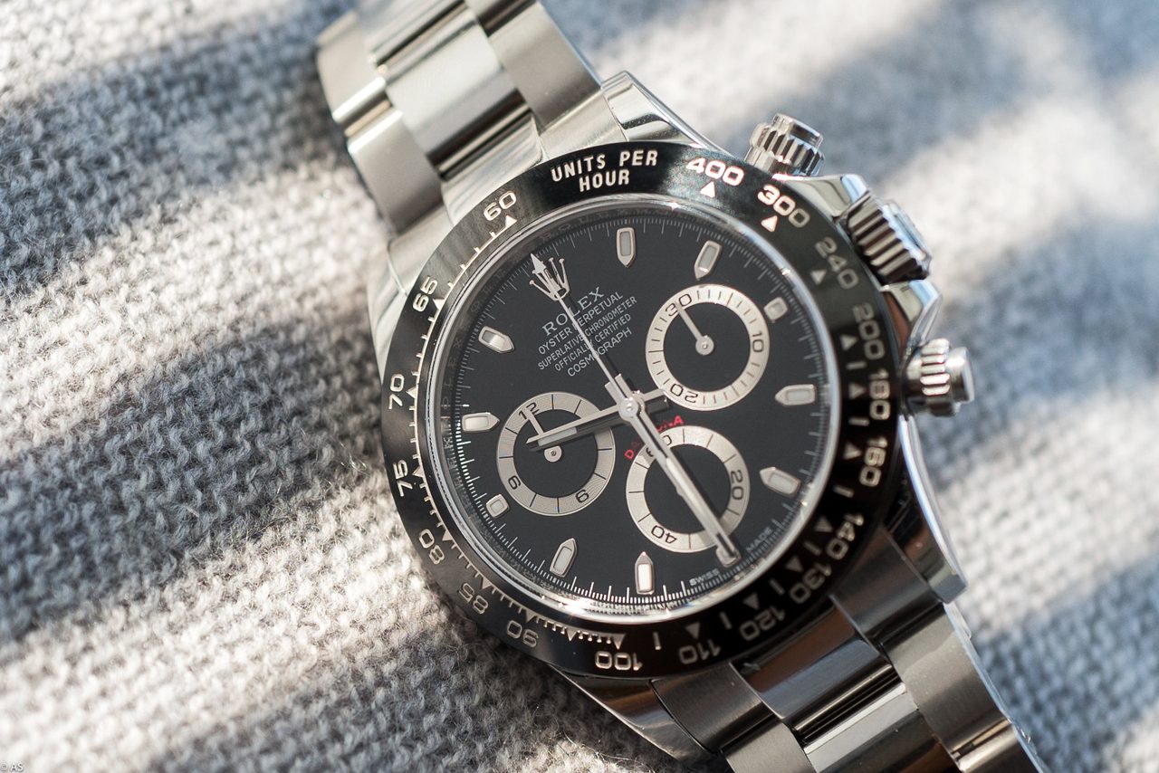 Stahl-Daytona mit schwarzem Zifferblatt, drei hellen Hilfszifferblättern und schwarzer Tachymeter-Lünette, schräg von oben auf einem grau-weiss gestreiften Wollstoff im Seitenlicht.