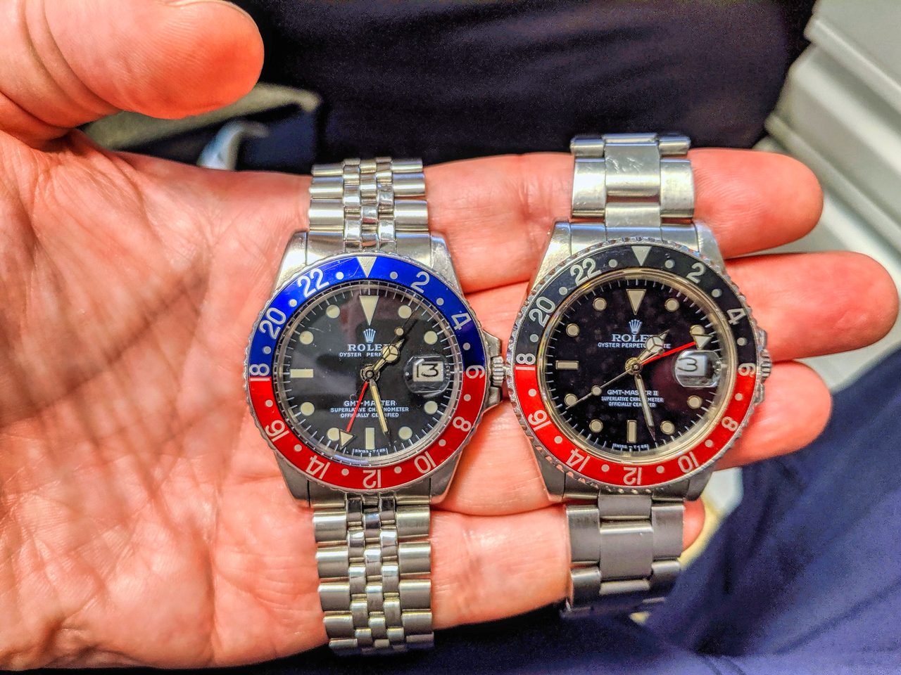 Zwei stählerne Rolex GMT-Master, links eine GMT-Master, rechts eine GMT-Master II, beide mit blau-roter Drehlünette, auf einer offenen Handfläche