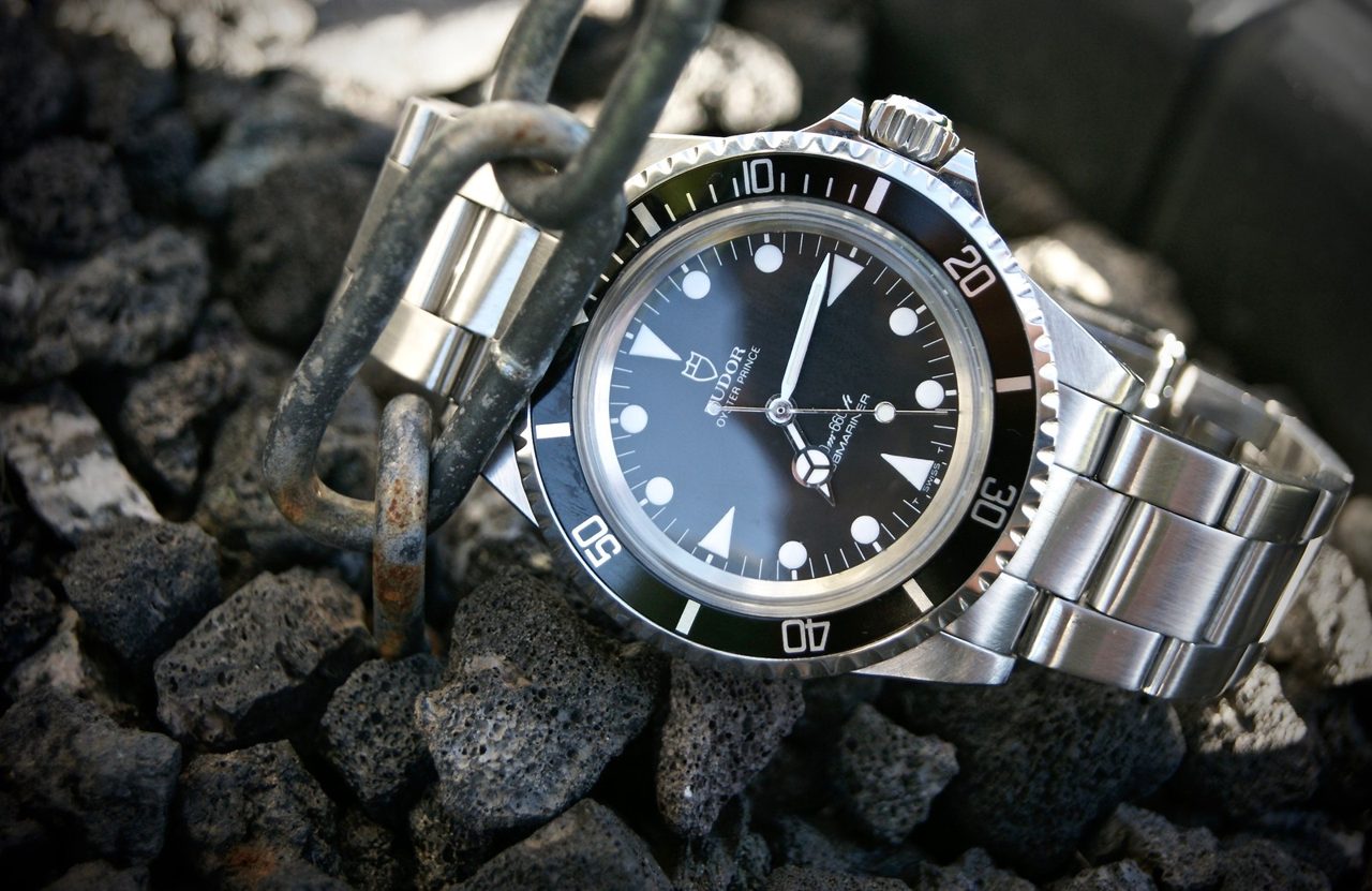 Tudor Oyster Prince Submariner mit schwarzem Zifferblatt, Schneeflocken-Zeiger und Stahlband, schräg auf dunklem Lavagestein neben einer rostigen Kette.